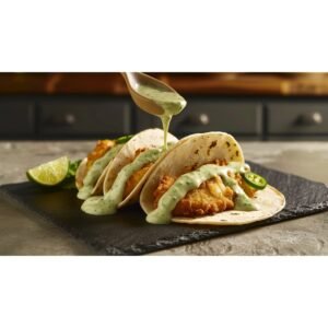 Creamy pale green jalapeño taco sauce drizzled over fish tacos on dark slate with lime wedge in warm home kitchen setting