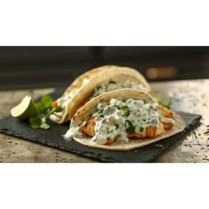 Cool creamy fish taco sauce drizzled over two crispy fish tacos with cilantro and lime on dark slate in warm natural kitchen light