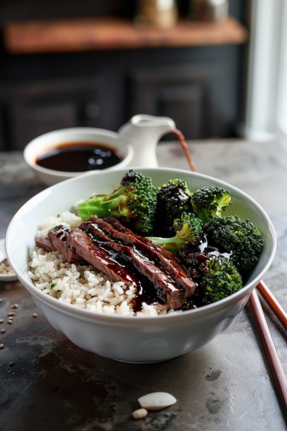 Oyster sauce broccoli beef rice bowl with dark glossy sauce ramekin and chopsticks on stone counter
