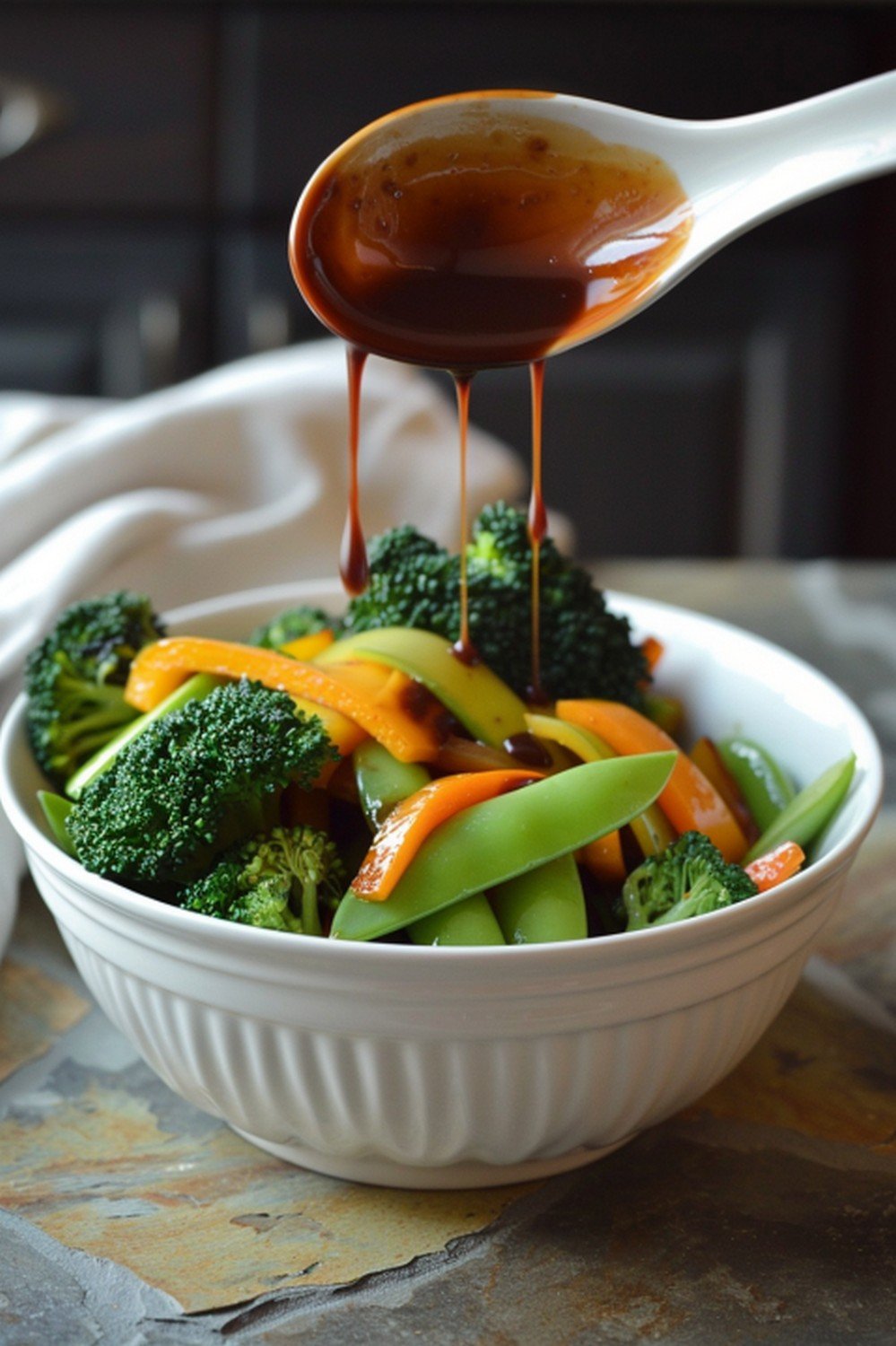 Vegetable stir fry sauce drizzled from ceramic spoon over colorful vegetables in white bowl on stone counter
